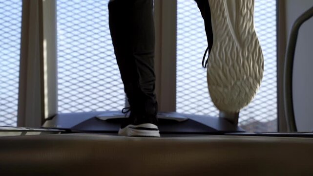 Person exercises on treadmill indoors. Feet move rhythmically, focused, steady. Background shows modern window with grid pattern. Bright daylight filters through the glass. Ideal for fitness, wellness