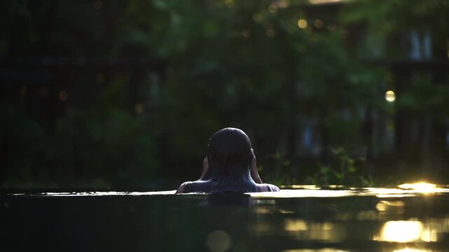 Dark water reflects golden light. Person emerges slowly from the surface. They raise their hands, soaking in warmth. Stillness holds the moment, tranquil and calm. Ideal for serene spa, wellness