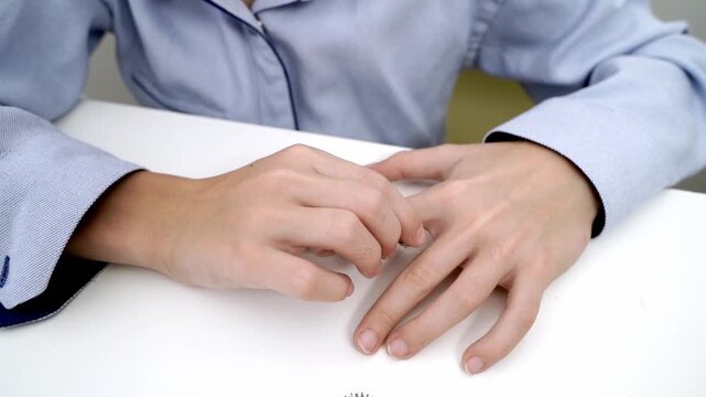 Person in blue shirt sits at white table. Hands fidget nervously over table surface. Focus on fingers, showing subtle anxiety or stress. Ideal for business, meeting, or personal pressure scenes