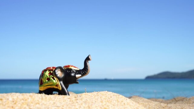 Tiny elephant figurine rests on warm sand. Blue sea stretches behind, calm and wide. Clear sky above, soft and serene. Sunlight glows gently on beach scene. Perfect for travel, vacation