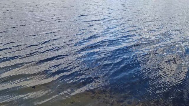 Blue water surface with light waves and reflections of the sky. Texture of water of a lake or river in daylight. The surface of the water is covered with small waves, in which the sky and clouds are r