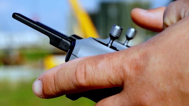 Hand grips remote control tightly. Thumb adjusts a dial with focused intent. Background hints at open field. Distant yellow structure suggests industrial zone. Steady grip implies concentration