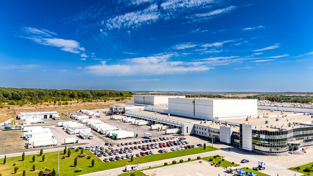Aerial view of modern industrial warehouse and logistics center. Wide aerial shot of a white industrial building with parking lots and trucks under a blue sky.