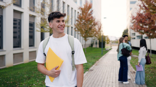 Young man smiling, holding folder, walking outside university building. Other students socializing in background