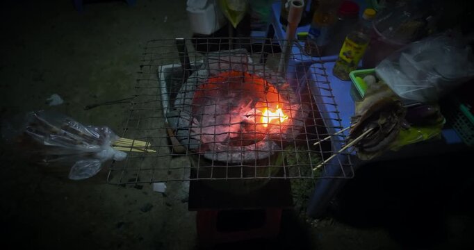 Slow motion close up of a traditional charcoal stove with a metal grill on top, featuring bright orange flames and smoke, set up for street food grilling at night