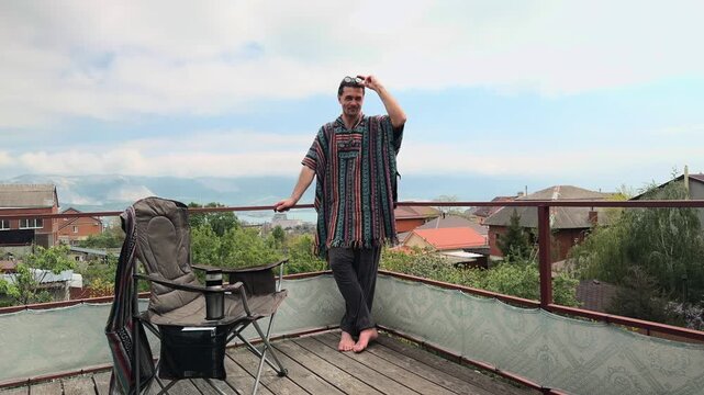 Authentic Bohemian Man with Ponytail wearing Colorful Ethnic South American Poncho and Round Sunglasses standing on Wooden Terrace Railing lifting Glasses and Smiling over Scenic Harbor Bay Vista