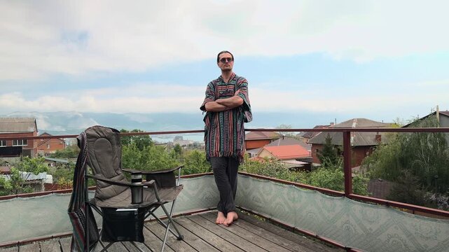 Creative Freelancer with Ponytail and Round Sunglasses wearing Ethnic Mexican Poncho standing on Balcony Crossing Arms and Looking at Camera with Scenic Harbor Mountain Bay Landscape Wide Shot