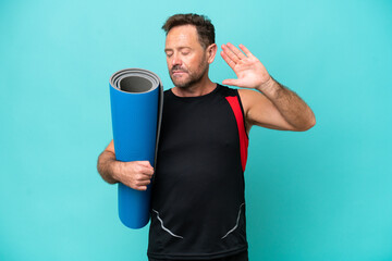 Middle age sport man going to yoga classes while holding a mat isolated on blue background making...