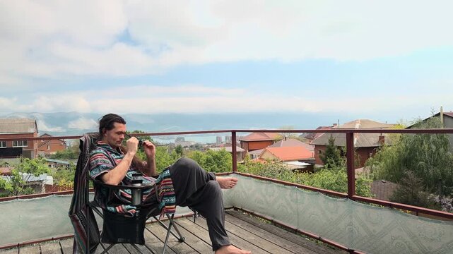 Serious Bohemian Man with Ponytail wearing Colorful Ethnic Poncho Sitting in Folding Chair on Terrace Taking Off Sunglasses and Thinking at Coastal Harbor Mountain View Wide Static Profile Shot