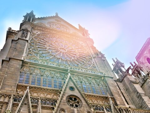 A dramatic bottom-up perspective of the iconic South Rose Window of Notre-Dame de Paris Cathedral. The Gothic stone tracery and stained glass are highlighted by a warm, ethereal lens flare against a s