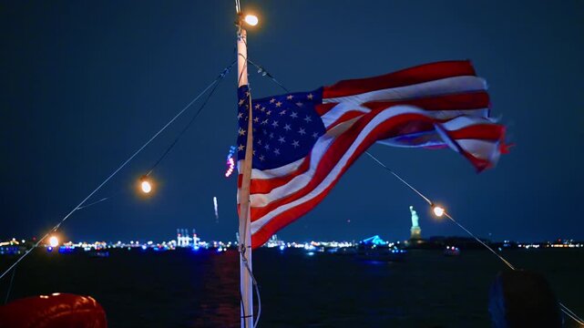 Wind flaps American flag on the boat mast at night. Fireworks and Statue of Liberty in the backdrop.