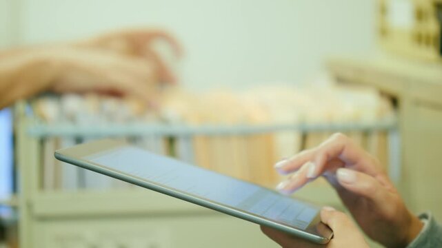 Professional woman using tablet computer to manage digital records in modern office while coworker searches physical files.