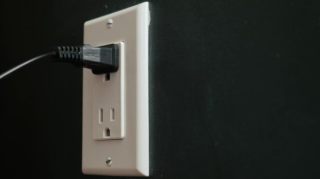Close up of hand plugging a black electrical cord into a white wall socket.