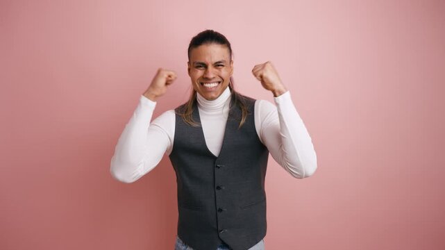 Cheerful hispanic man with long hair clenching his fists and celebrating victory. Enthusiastic guy screaming with joy in studio