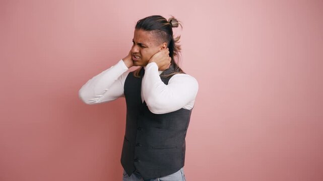 Unwell man massaging his stiff neck and grimacing from intense pain. A tired person feeling muscle ache and tension against a pink background