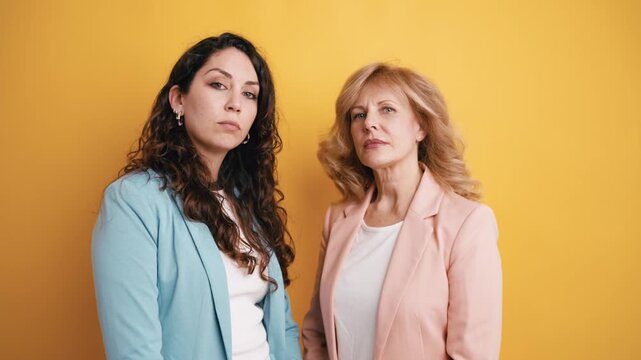 Two professional women, a mother and daughter or business partners, standing with confidence against a yellow background in a studio setting
