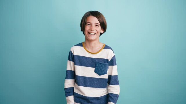 Portrait of a happy preteen boy with long hair smiling and laughing. Cheerful kid expressing positive emotions against a blue background