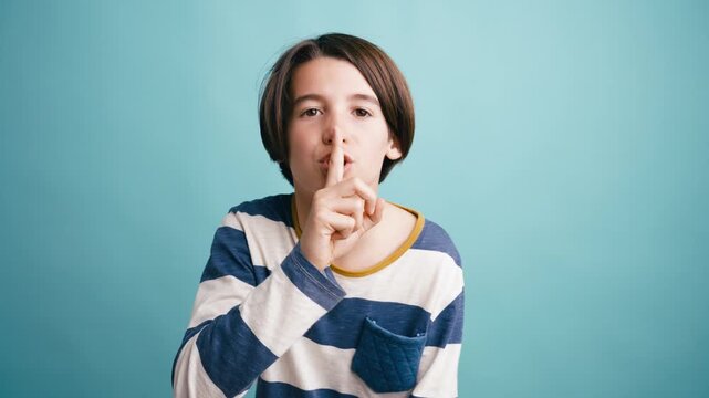 Young boy putting his finger on his lips, asking for silence. The studio portrait captures a confidential and quiet moment against a blue background