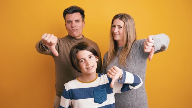 Dissatisfied family of three showing a thumbs down gesture. Unhappy parents and their son expressing disapproval on a yellow background
