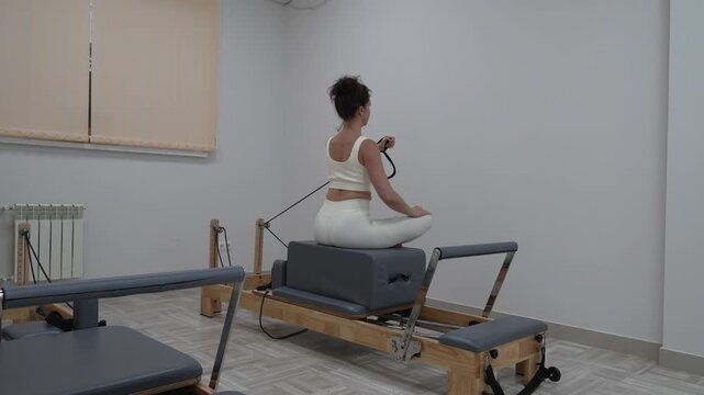 Pilates reformer workout woman in white activewear performs arm exercises while sitting on a reformer machine in a bright studio