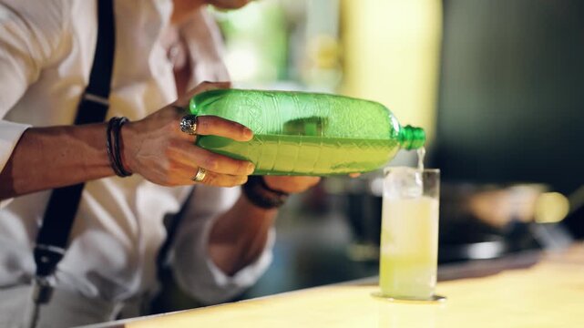 Professional bartender pouring a fresh, fizzy drink from a green bottle into a glass with ice cubes, making a non alcoholic cocktail