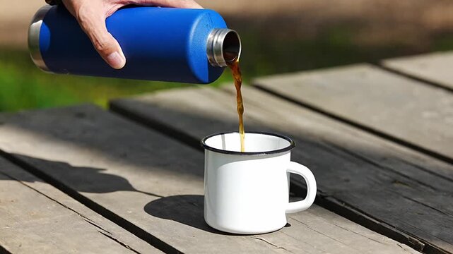 Pouring hot coffee from a blue thermos into an enamel mug
