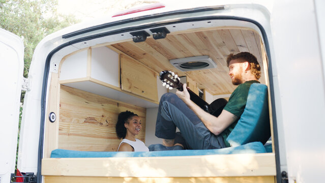 Couple relaxing inside a camper van, man playing acoustic guitar and woman listening happily during a road trip adventure