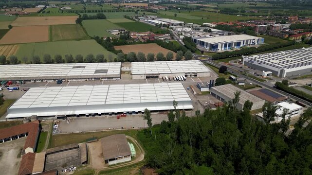 Aerial View of Industrial Park: An expansive industrial park is captured from an aerial perspective, showcasing rows of large warehouses, agricultural fields, and a network of roads under a hazy sky.