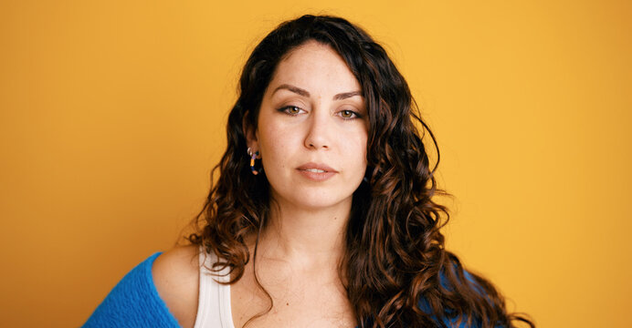 Confident young woman with curly brunette hair and neutral expression, direct gaze in a blue sweater against vibrant yellow background