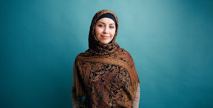 Woman wearing a patterned headscarf and modest clothing, standing confidently against a blue background, looking forward