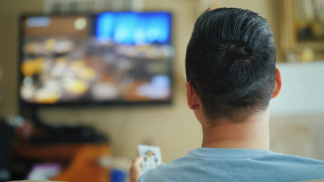 Young man in living room holding remote control and switching channels on television screen.
