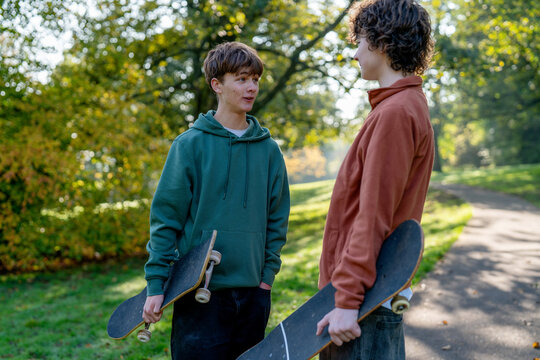 Two young people talk while holding skateboards in a park during daytime
