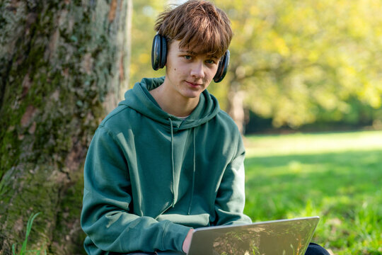 Teen listens to music on headphones while using a laptop under a tree in a park during a sunny afternoon