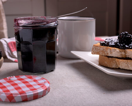 Toasted bread with homemade blackberry jam on a white plate served with a cup of tea, fresh berries and a glass jar over a neutral stone background