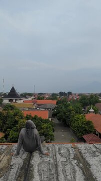 An unrecognisable person sits on an abandoned building structure, gazing at the cityscape under a vast cloudy sky. The view features rooftops, trees, and a prominent mosque in the distance.	