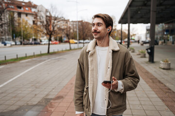 A stylish young male with a mustache walks outdoors on a sidewalk, smiling while holding a smartphone and listening to music through wired earphones during a casual city commute. © Dusko