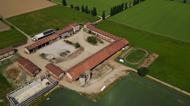 Agricultural Estate Aerial View: An expansive agricultural estate, featuring a cluster of farm buildings with rustic red roofs, surrounded by vibrant green fields and farmland. Settala, Lombardy