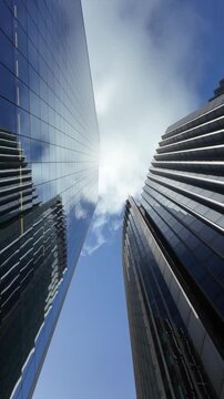 Stunning low-angle perspective of towering glass skyscrapers reflecting a beautiful blue sky with fluffy clouds as the view pans up towards a bright, sunny day in a major financial district
