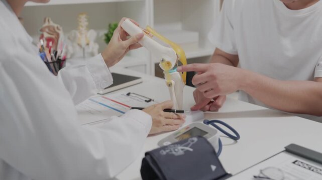 A young man sat in an examination chair, complaining of knee pain, while a female doctor recorded his symptoms in his medical history during a physical examination with a knee bone model on the table.