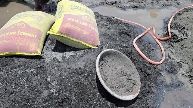 A construction scene: yellow cement bags on dark gravel. A plastic mortar pan for transporting sand sits nearby with wet mortar, next to an orange hose in a muddy area. Essential tool.