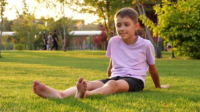 Happy boy relaxing on grass in park at sunset. Child enjoys nature and smiles. Kids play in background, creating joyful atmosphere. Perfect for family and outdoor themes.