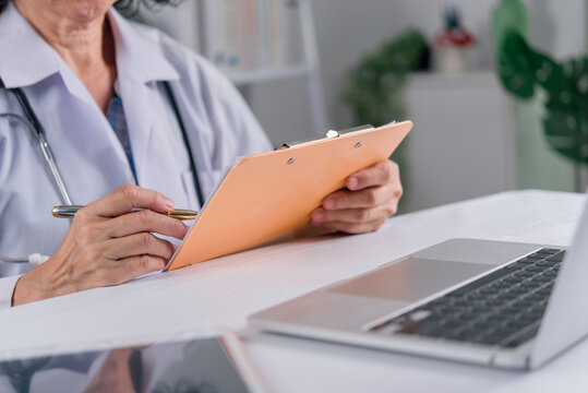 Senior female doctor holding clipboard and pen working with laptop in hospital
