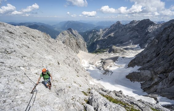 Mountaineer with helmet in a secured via ferrata, Zugspitze via ferrata, ascent to the Zugspitze, view of mountain basin with snowfield of the H&ouml;llentalferner and H&ouml;llental, Wetterstein range, Wetterstein range, Bavaria, Germany