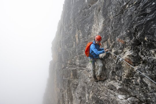 Mountaineers on a steep rock face on the Via Ferrata Oliva Detassis via ferrata in fog, steep mountains covered in clouds, Brenta Mountains, Trentino, Italy