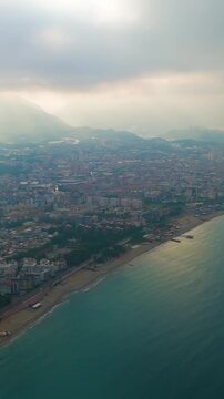 Vertical video. Alanya, Turkey. Aerial drone view of the Mediterranean coastline stretching past Oba Beach and Tosmur Beach, with sun rays breaking through low clouds over the Taurus Mountains and tu