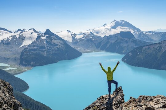 Young man standing on a rock, stretching his arms in the air, view of mountains and glacier with lake Garibaldi Lake, peaks Panorama Ridge, Guard Mountain and Deception Peak, Garibaldi Provincial Park, British Columbia, Canada