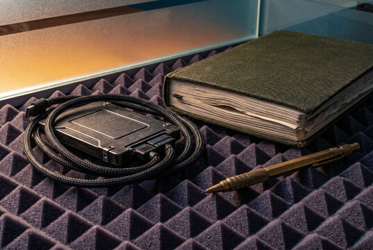 Rugged solid-state drive and a weathered canvas notebook resting near an azure glass partition. Raking light emphasizes heavy braided cables and deep architectural shadows.
