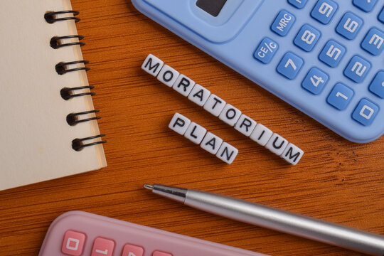 Letter blocks spelling moratorium plan with calculators, notebook, and pen representing financial relief, debt suspension, budgeting, and economic recovery concept.