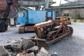 Fototapeta premium Rusted crawler tractor with front blade stands beside large mining machinery and broken stones on a gravel yard. The scene represents quarry equipment, construction history, mechanical decay, heavy in
