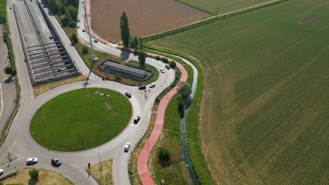 Roundabout Highway Interchange: An aerial view showcases a modern highway interchange with a large roundabout, surrounded by agricultural fields and a tree-lined road. Pantigliate, Milan, Italy